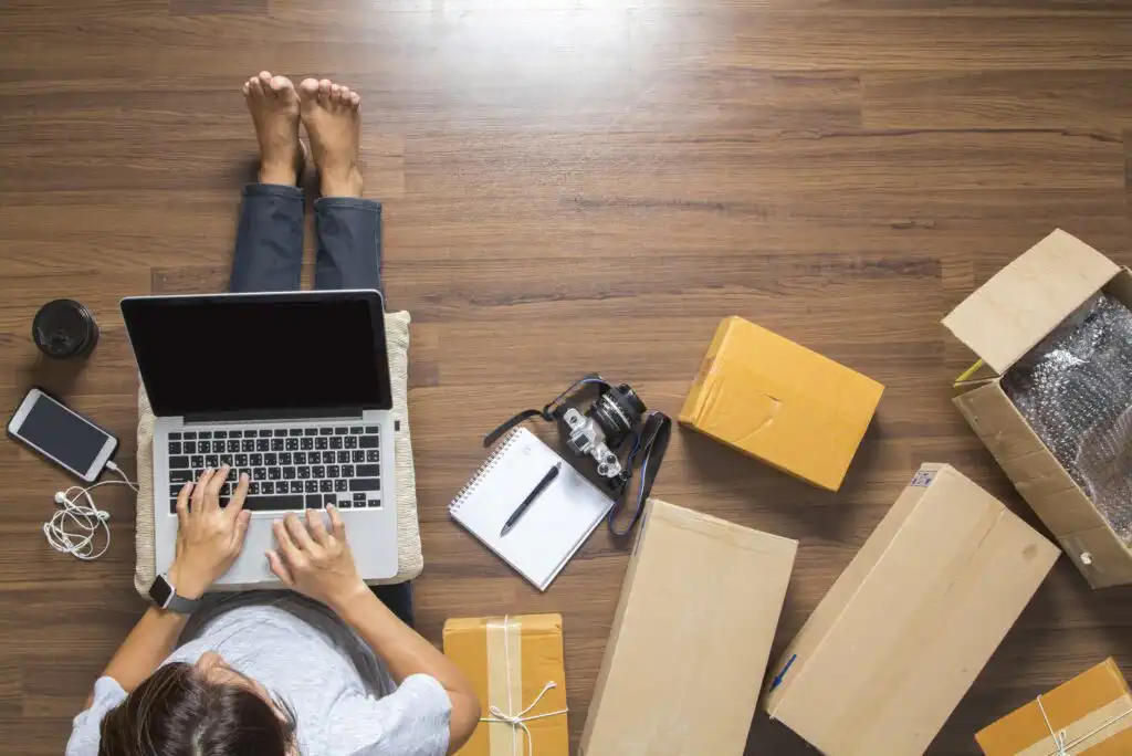 A person sits on a wooden floor using a laptop, surrounded by cardboard boxes, a notebook with a pen, a camera, a cup of coffee, a phone, and earphones. The scene suggests online business or working from home.