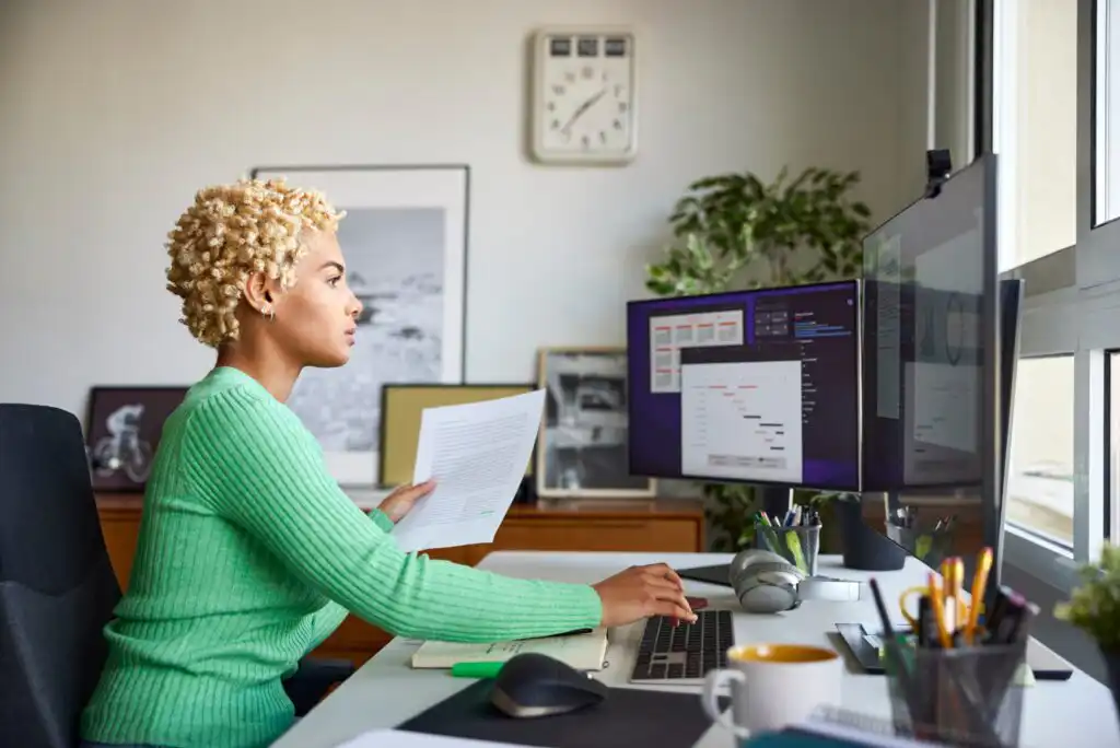 A person with short curly blonde hair, wearing a green sweater, sits at a desk working on a computer with two monitors. They hold a document and look focused. The desk has office supplies and papers, and there’s a clock on the wall.