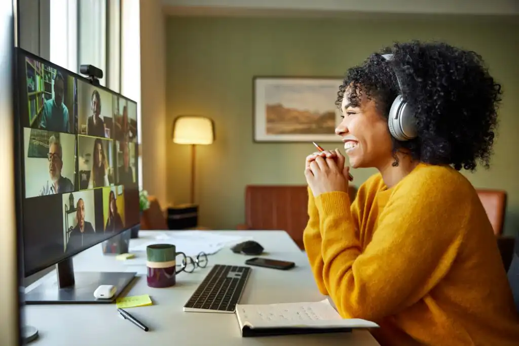 A woman wearing headphones and a yellow sweater smiles at her computer during a video conference call, with several people visible on the screen. She sits at a desk with a notebook, keyboard, and mug.