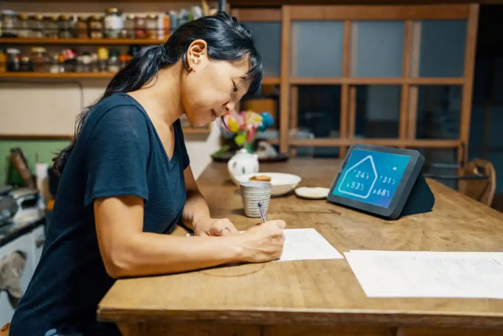 A woman sits at a wooden kitchen table, smiling as she writes on papers. A tablet displaying graphs and numbers is propped up nearby, and kitchen shelves with jars are visible in the background.
