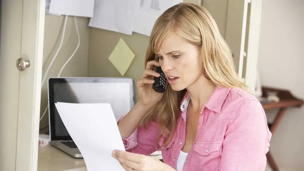 A woman wearing a pink shirt is sitting at a desk, holding a phone to her ear and looking concerned while reading a document. A laptop and papers are visible in the background.