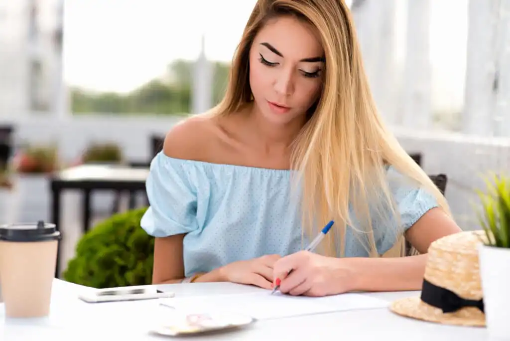 A young woman with long blonde hair and an off-shoulder blue top sits at an outdoor table, writing on paper with a pen. A coffee cup, phone, and straw hat are on the table in front of her.