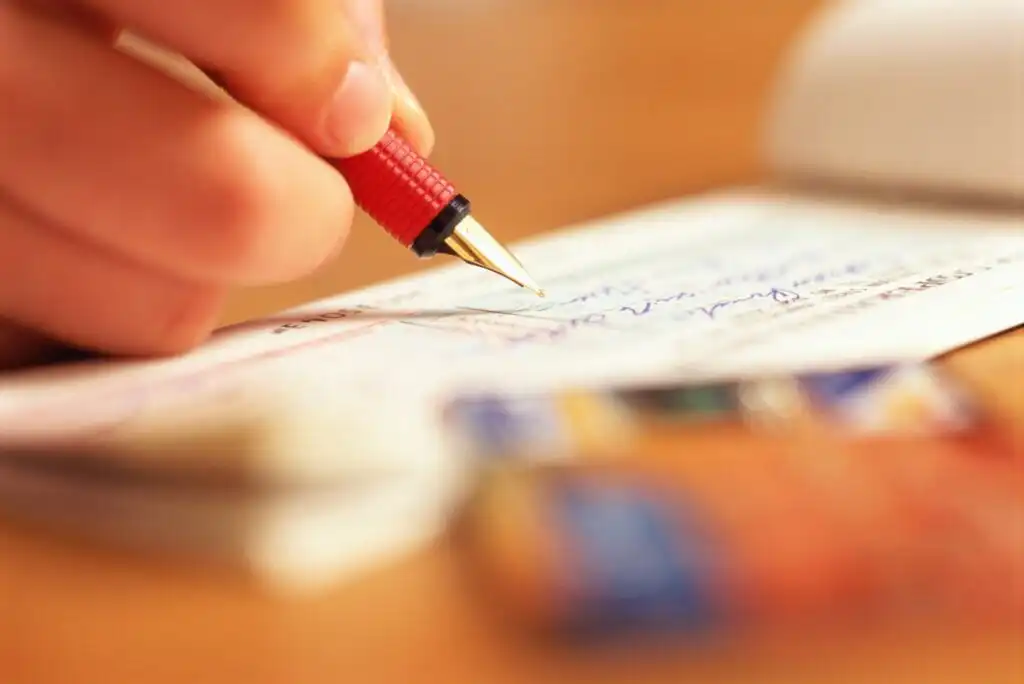 A close-up of a hand holding a red fountain pen, writing on a check. The background is blurred, showing indistinct objects, possibly credit cards, and a soft, warm lighting.