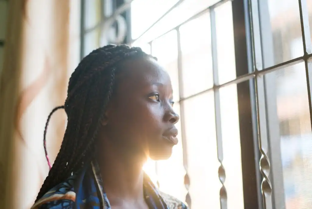 A woman with braided hair gazes thoughtfully out of a window with metal bars, sunlight shining on her face from behind.