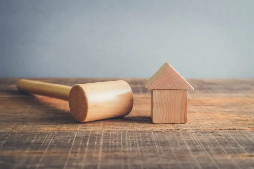 A wooden gavel lies on a rustic wooden table next to a small house-shaped block, symbolizing real estate law or property auction.