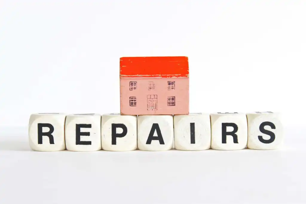 A small model house with a red roof sits atop wooden blocks that spell out REPAIRS against a white background.
