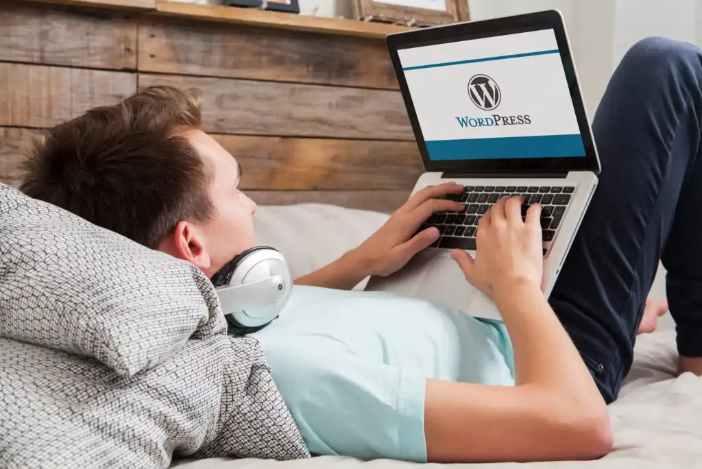 A person lying on a bed, wearing headphones around their neck, using a laptop that displays the WordPress logo on the screen.