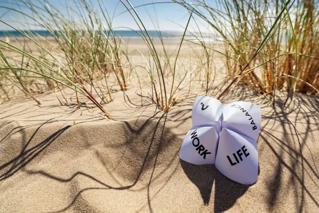 A paper fortune teller with the words WORK, LIFE, BALANCE, and ? sits on sandy beach among tall grass, with the ocean and blue sky in the background.