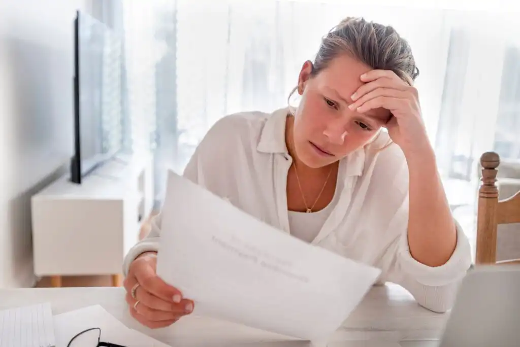 A woman sits at a desk, looking worried as she reads a document. She holds her forehead with one hand and holds the paper with the other, surrounded by papers, a pen, and a laptop.