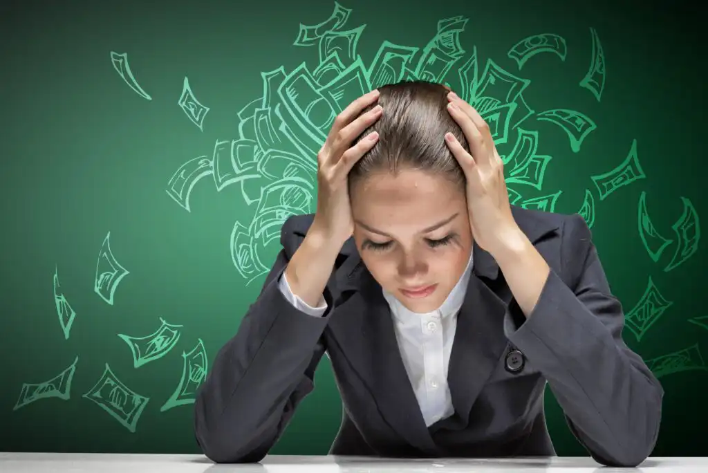 A woman in a business suit sits at a desk with her head in her hands, looking stressed. Illustrated dollar bills surround her head against a green background, symbolizing financial stress or worry.