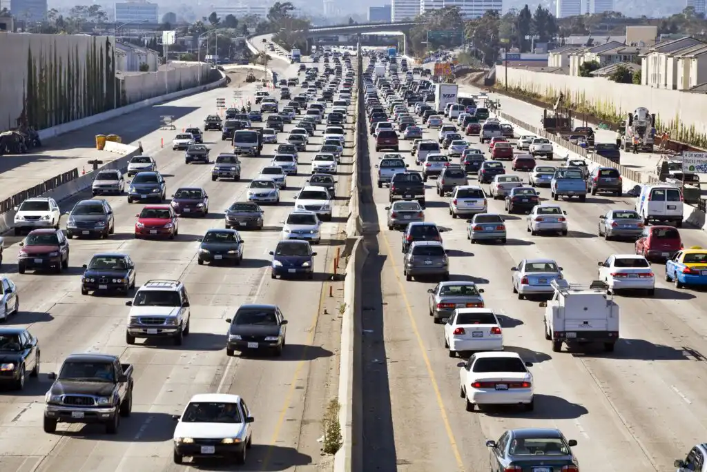 Heavy traffic on a multi-lane freeway, with cars, trucks, and SUVs filling both directions. The scene is urban, with buildings and trees visible on both sides of the road.