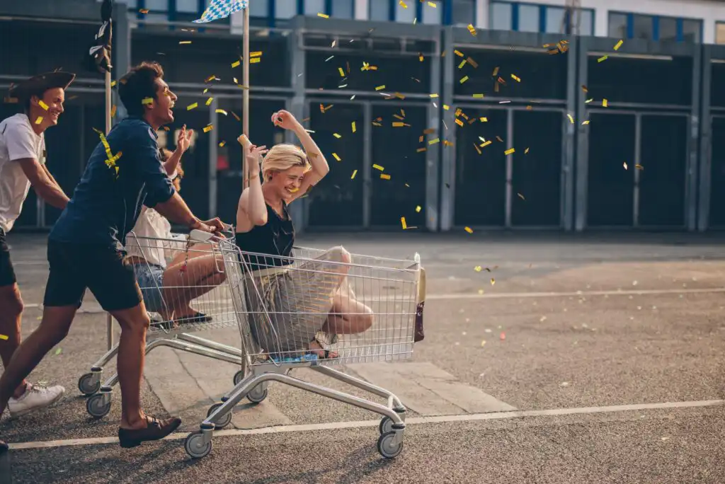 Four young adults have fun outdoors; two push a shopping cart with a laughing woman inside while confetti flies around them, all on a sunny day in an empty parking lot.