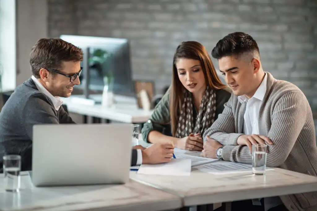 Three people sit at a table in an office, reviewing documents together. A man in glasses points at a paper while a woman and another man listen attentively. A laptop and glasses of water are on the table.