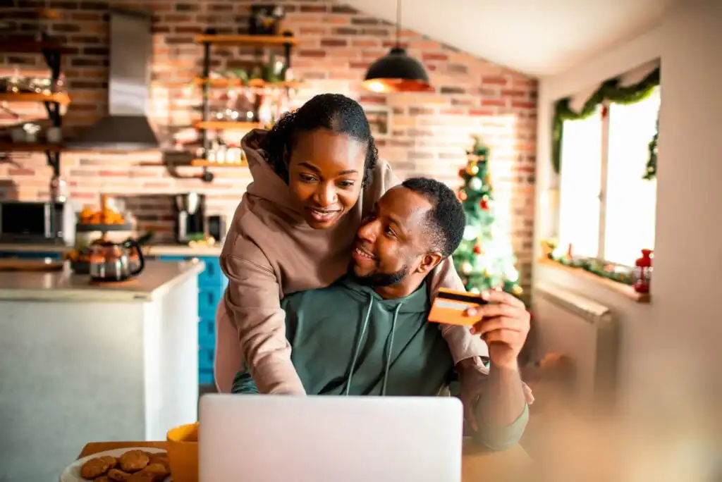 A smiling couple sits in a cozy kitchen decorated for the holidays. The woman hugs the man from behind as he holds a credit card and they look at a laptop together, appearing to shop online.