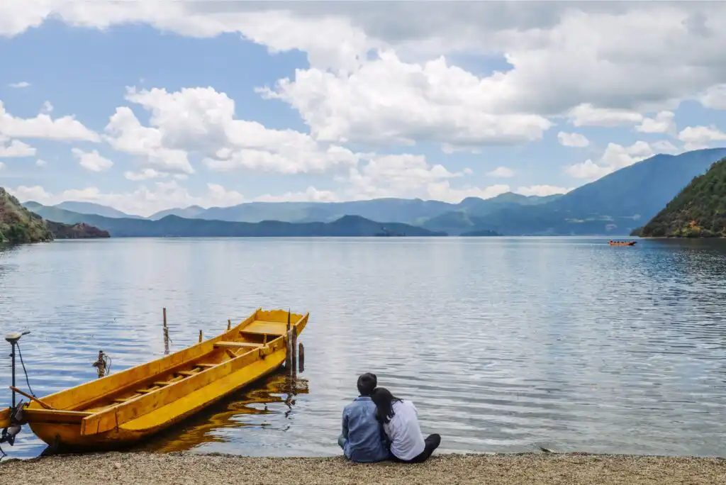 A couple sits on a pebbled shore facing a calm lake, with a yellow wooden boat nearby and mountains under a partly cloudy sky in the background.