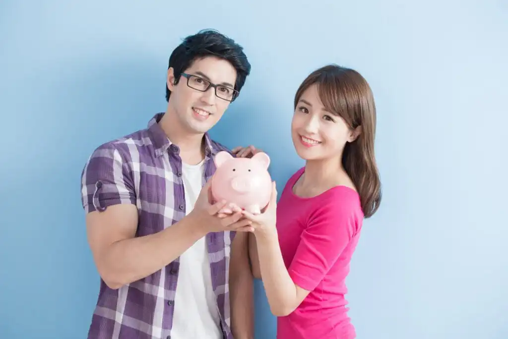A smiling man and woman stand together against a light blue background, both holding a pink piggy bank in front of them, suggesting the concept of saving money or financial planning.
