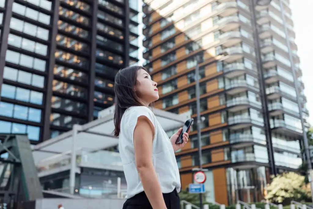 A woman stands outdoors in a city, holding a phone and looking up at tall modern office buildings with glass and metal facades.