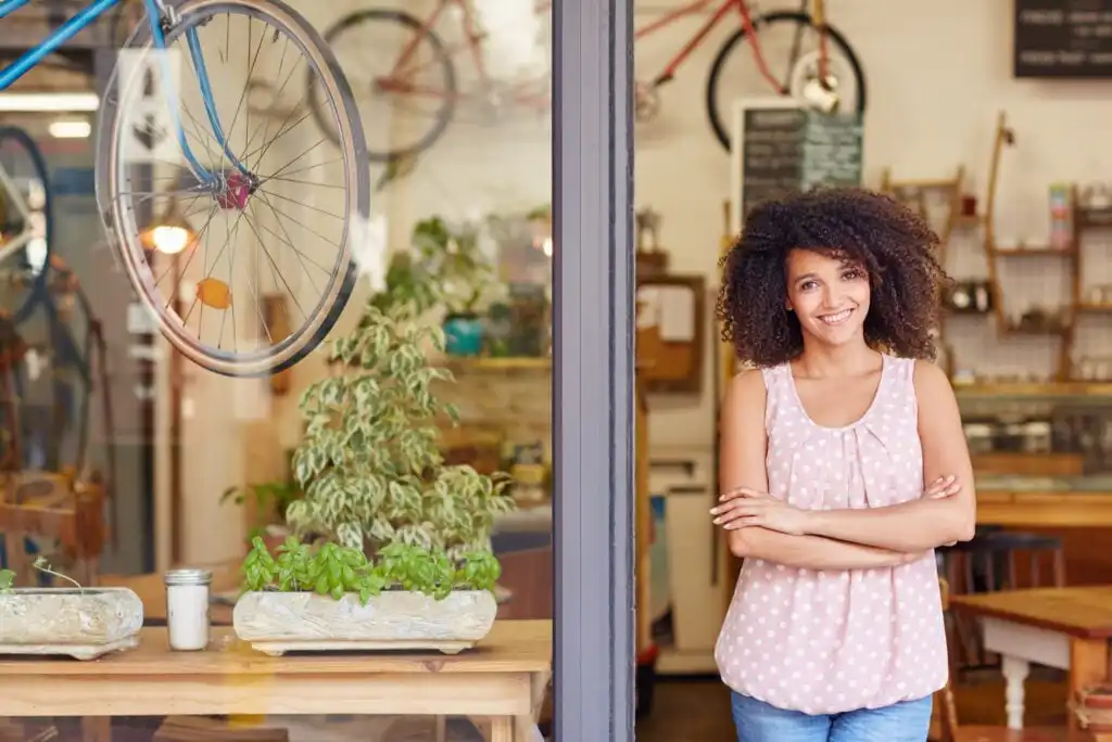 A woman with curly hair wearing a pink polka-dot top stands smiling with arms crossed in the doorway of a cozy shop. Bicycles, plants, and shelves are visible in the inviting interior behind her.