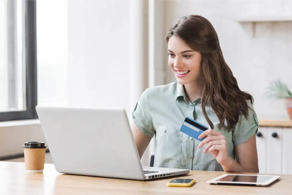 A woman sits at a desk smiling at her laptop screen while holding a credit card. A coffee cup, smartphone, and tablet are also on the desk in a bright, modern room.