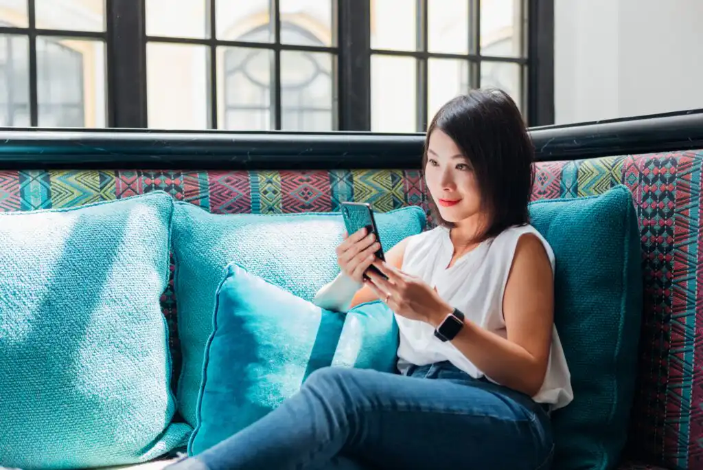 A woman sits on a colorful couch with teal cushions, looking at her smartphone and smiling. She wears a white sleeveless top, blue jeans, and a smartwatch, with large windows in the background.
