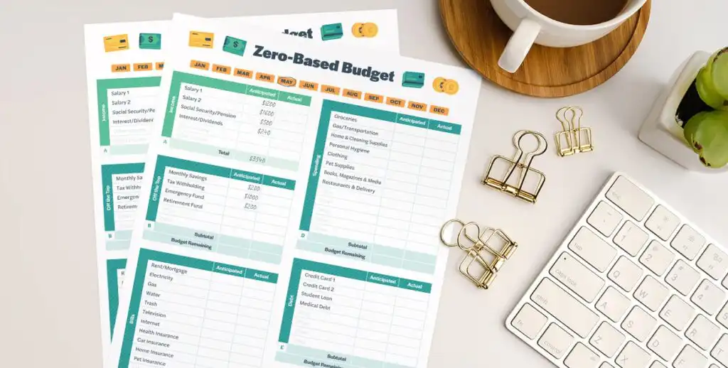 Three printed zero-based budget sheets are on a desk next to a keyboard, a cup of coffee on a coaster, two gold binder clips, and a small bowl of green apples. The budget sheets list categories and amounts.